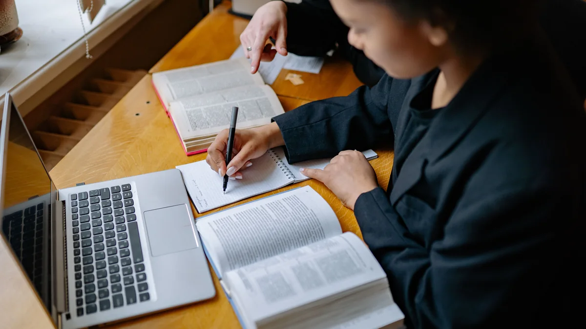 Estudantes fazem anotações com livros e notebook sobre a mesa.
