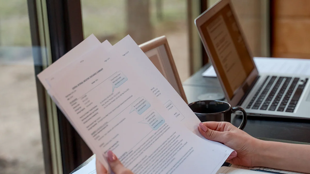 Pessoa revisa documentos diante de notebooks em uma mesa.